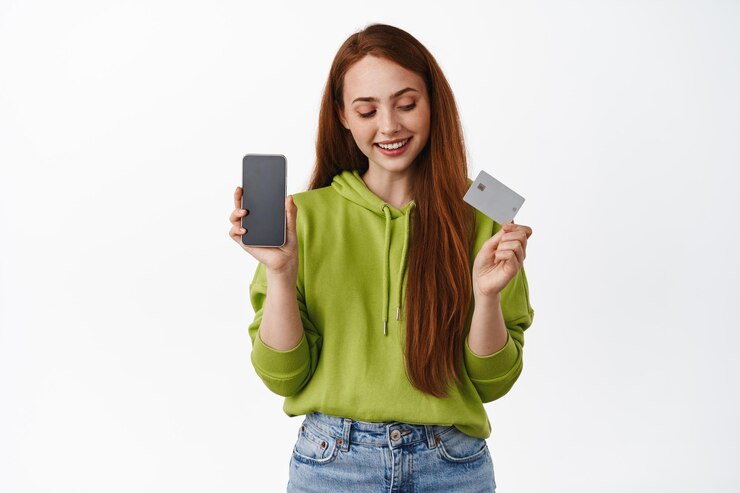 Foto grátis jovem ruiva com sorriso branco, olhando para o cartão de crédito, mostrando a tela vazia do aplicativo do telefone, introduzindo uma nova interface, aplicativo no celular, fundo branco.