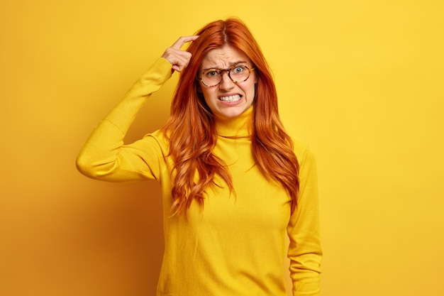 Jovem ruiva decepcionada tem dúvidas, coça a cabeça e cerra os dentes, usa gola olímpica casual, tem memória ruim.