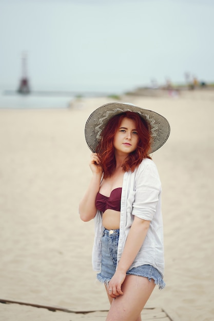 Jovem ruiva em um grande chapéu redondo e camisa branca em pé na areia na praia