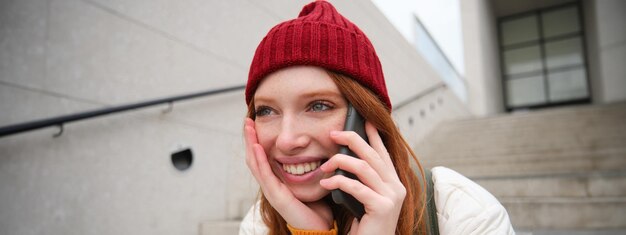 Foto grátis jovem ruiva estilosa de chapéu vermelho senta-se na rua e fala no celular tem telefone