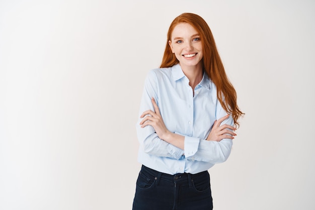 Jovem ruiva feliz sorrindo para a frente, cruzando os braços no peito, confiante, em pé na blusa do escritório sobre a parede branca