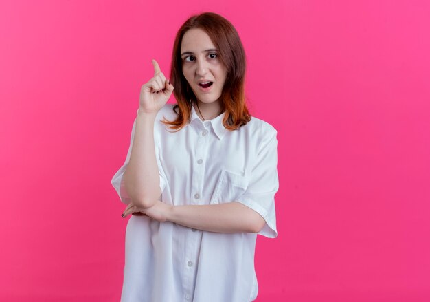 Jovem ruiva impressionada apontando para cima isolado em um fundo rosa