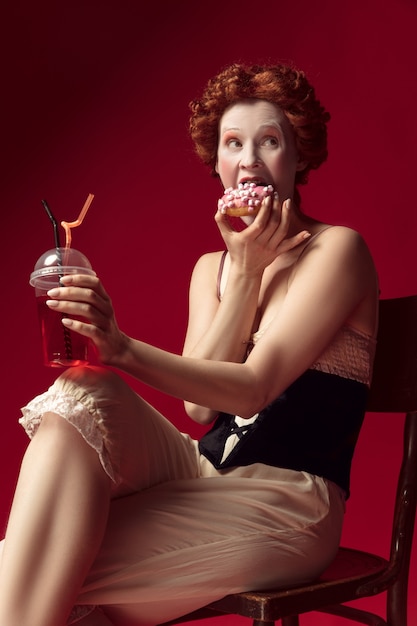 Foto grátis jovem ruiva medieval como uma duquesa com espartilho preto e roupas de dormir, sentada em uma cadeira no espaço vermelho com uma bebida e donut