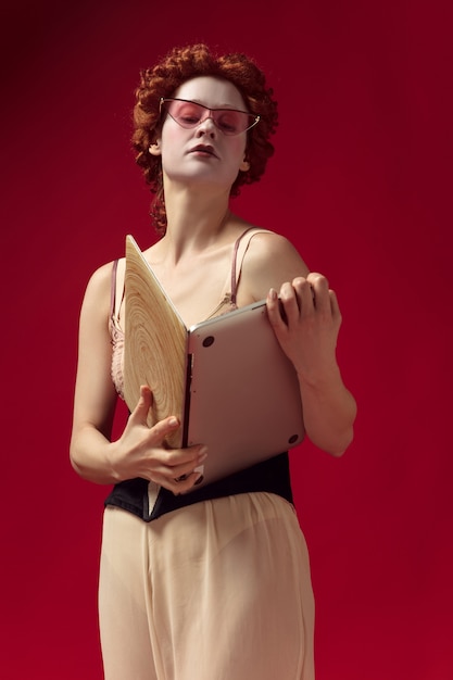 Jovem ruiva medieval como uma duquesa em espartilho preto, óculos escuros e roupas de noite em pé na parede vermelha com um laptop como um livro. conceito de comparação de eras, modernidade e renascimento.