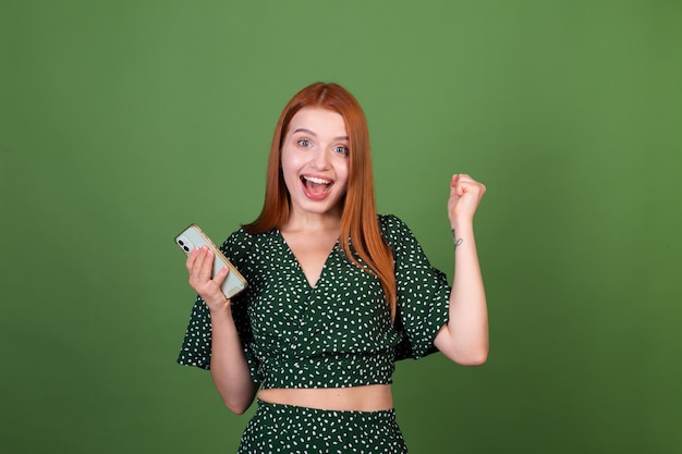 Foto grátis jovem ruiva na parede verde com mensagens de texto no celular, conversando animada, mostrando gesto do vencedor