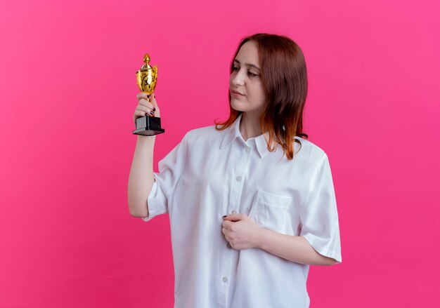 Foto grátis jovem ruiva segurando e olhando para a taça do vencedor isolada na rosa com espaço de cópia