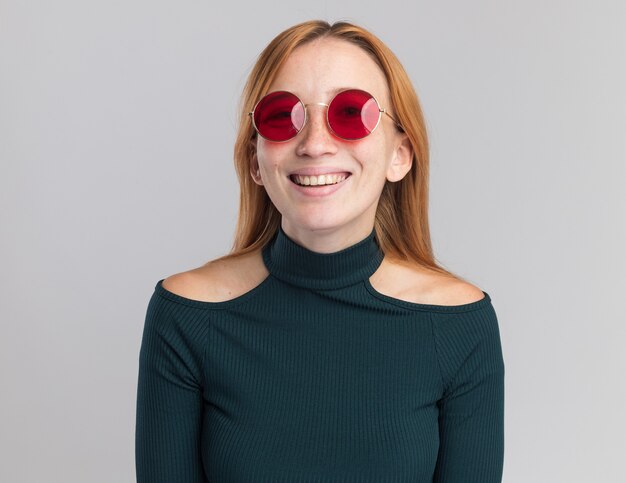 Jovem ruiva sorridente com sardas em óculos de sol olhando para a câmera em branco