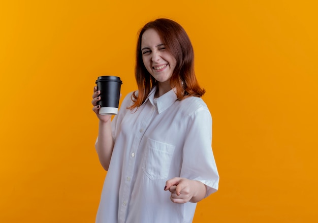 Jovem ruiva sorridente segurando uma xícara de café e mostrando um gesto isolado na parede amarela