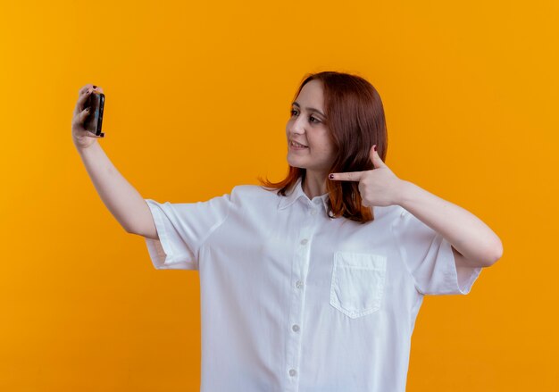 Jovem ruiva sorridente tira uma selfie e aponta para um telefone isolado em fundo amarelo