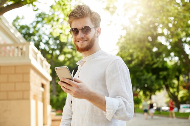 Jovem ruivo positivo e sorridente com barba e brinco em óculos de sol, olhando através de redes sociais em seu smartphone a caminho de casa.