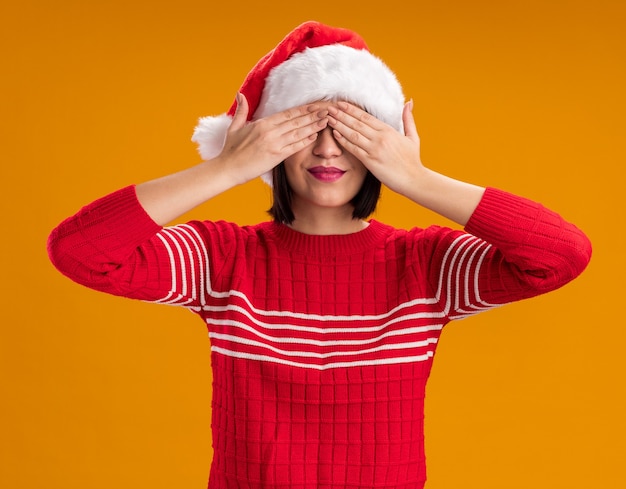 Foto grátis jovem satisfeita com chapéu de papai noel cobrindo os olhos com as mãos isoladas na parede laranja