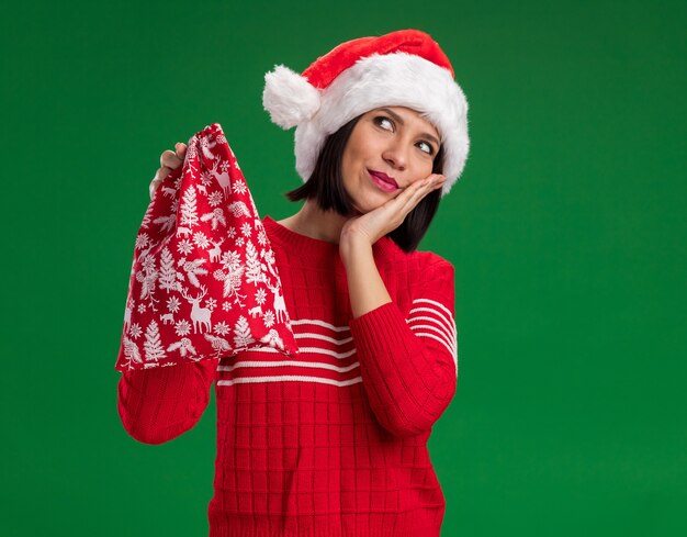 Jovem satisfeita com chapéu de Papai Noel segurando um saco de presente de Natal, mantendo a mão no rosto, olhando para cima, isolado em um fundo verde com espaço de cópia