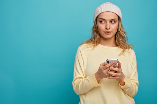 Jovem satisfeita com um chapéu de inverno segurando um celular e olhando para o lado