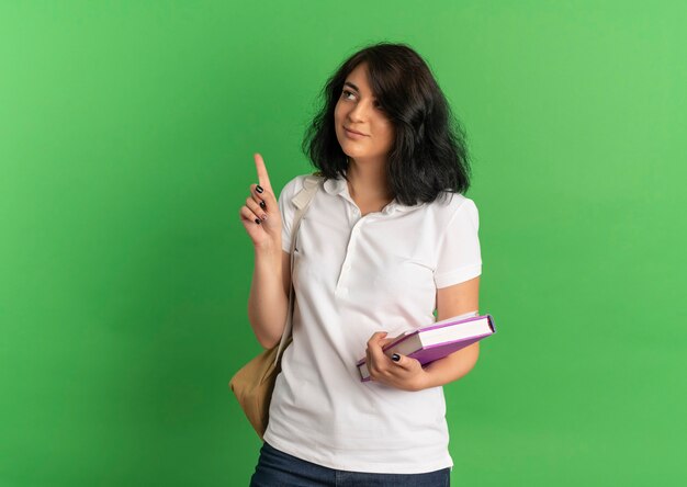 Jovem satisfeita com uma aluna bonita caucasiana vestindo uma bolsa atrás de olhares e apontando para cima segurando livros em verde com espaço de cópia