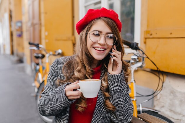 Jovem satisfeita com uma jaqueta cinza falando ao telefone e tomando café em um café de rua