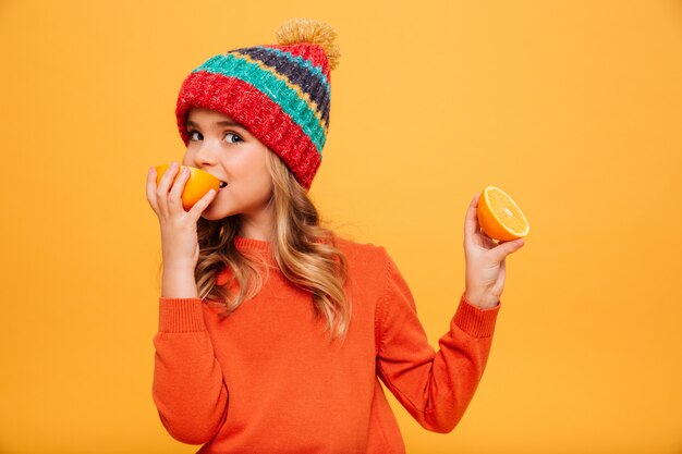 Jovem satisfeita de suéter e chapéu comendo laranja enquanto olha para a câmera sobre laranja