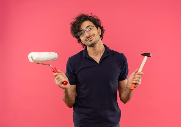 Jovem satisfeito com uma camisa preta e óculos ópticos segurando um rolo de pintura e um martelo isolados na parede rosa
