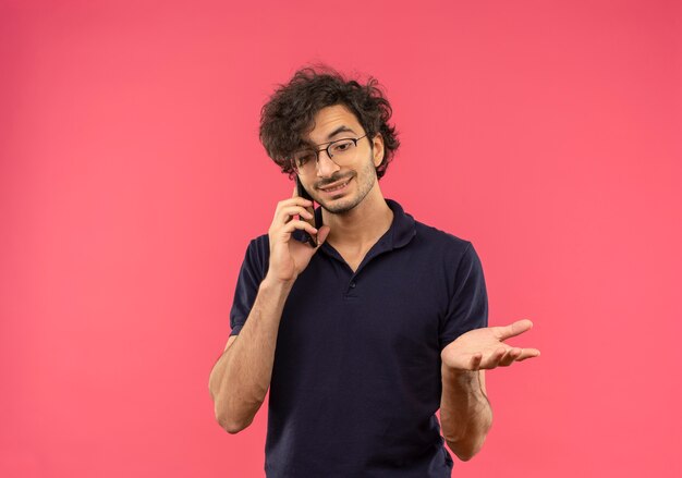 Jovem satisfeito de camisa preta com óculos óticos falando no telefone isolado na parede rosa