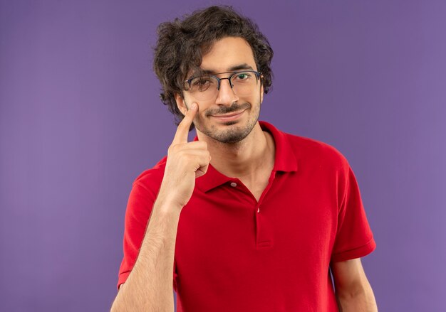 Jovem satisfeito de camisa vermelha com óculos ópticos apontando para um olho isolado na parede violeta
