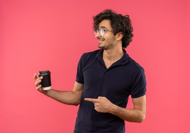 Jovem satisfeito em uma camisa preta com óculos ópticos segura e aponta para a xícara de café, olhando para o lado isolado na parede rosa