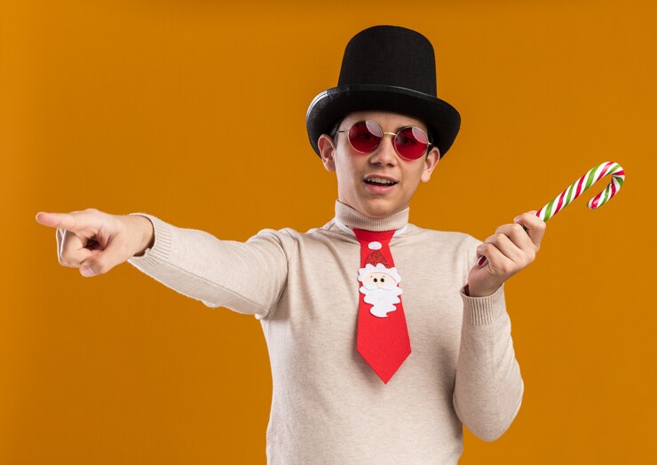 Foto grátis jovem satisfeito usando chapéu com gravata de natal e óculos segurando balas de natal ao lado isoladas em fundo amarelo