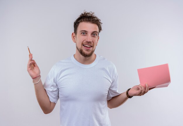 Jovem satisfeito vestindo camiseta branca levantando lápis e caderno na mão em fundo branco isolado