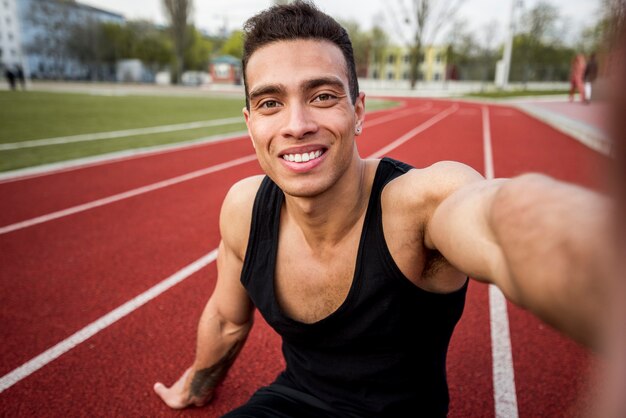 Jovem saudável fitness sentado na pista de corrida, tendo selfie no telemóvel