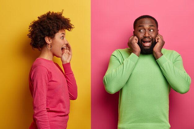 Jovem secreta fica de perfil, sussurra segredo para o namorado que não quer ouvi-la, tapa ouvidos, evita fofocar. Mulher afro-americana engraçada contando informações secretas e fazendo poses de lado