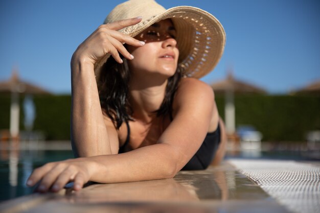 Jovem sedutora relaxante ao sol perto da piscina com um chapéu de palha em um dia ensolarado. Conceito de resort e verão.