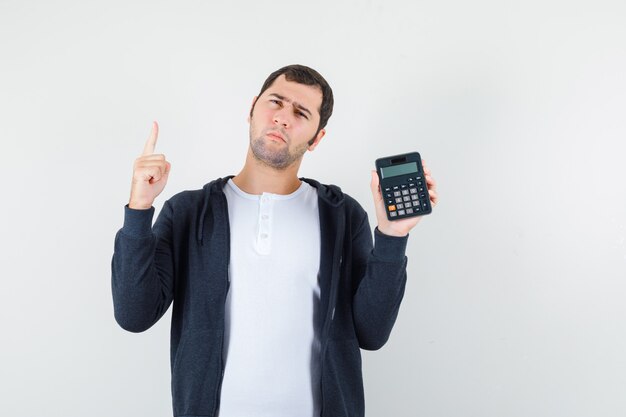 Jovem, segurando a calculadora e levantando o dedo indicador em gesto de eureka em t-shirt branca e capuz preto com zíper frontal e olhando pensativo. vista frontal.