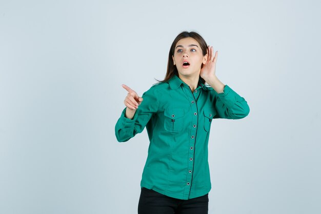 Jovem, segurando a mão perto do ouvido para ouvir algo, apontando para longe em blusa verde, calça preta e olhando focado. vista frontal.