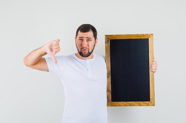 Jovem, segurando a placa preta enquanto mostra o polegar para baixo em uma camiseta branca e olhando descontente, vista frontal.