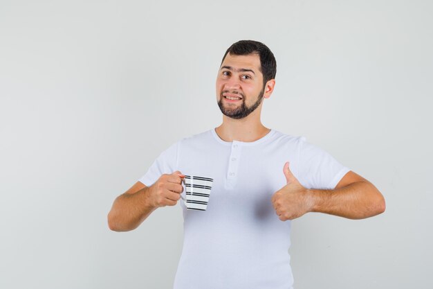 Jovem, segurando a xícara, aparecendo o polegar em uma camiseta branca e olhando otimista, vista frontal.