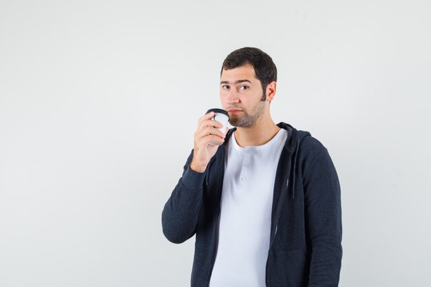 Jovem segurando a xícara de café para viagem em uma camiseta branca e um capuz preto com zíper na frente e olhando sério, vista frontal.