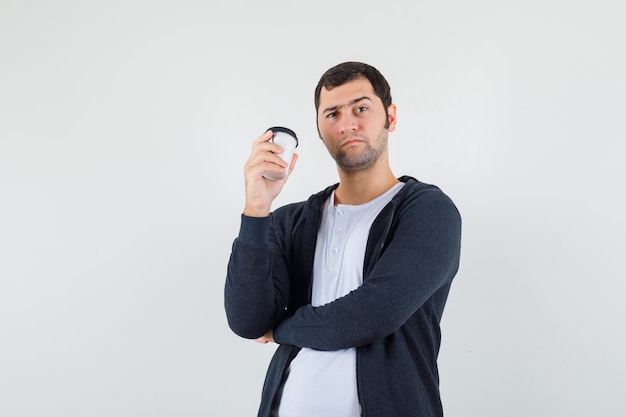 Jovem segurando a xícara de café para viagem em uma camiseta branca e um capuz preto com zíper na frente e olhando sério, vista frontal.