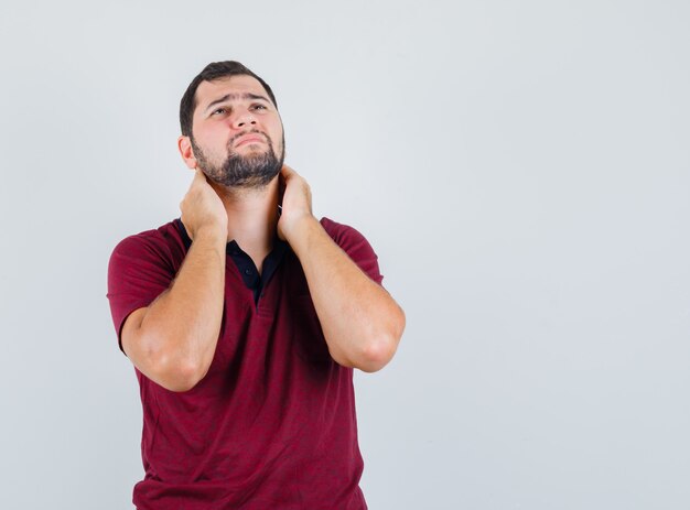 Jovem, segurando as mãos em seu pescoço em uma camiseta vermelha e parecendo descontente. vista frontal.