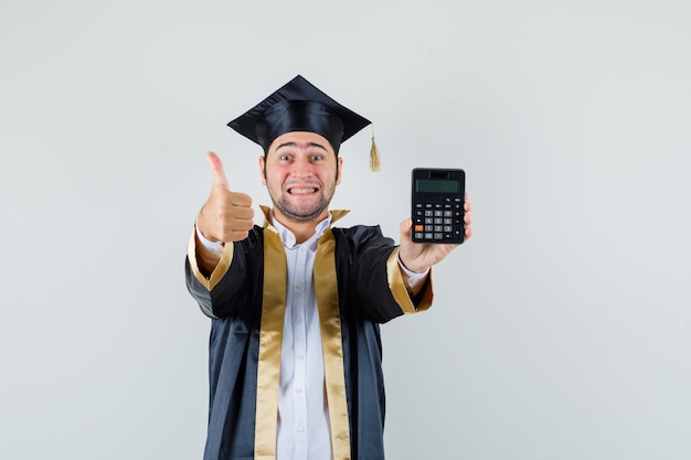 Jovem segurando calculadora, aparecendo o polegar em uniforme de pós-graduação e olhando alegre, vista frontal.