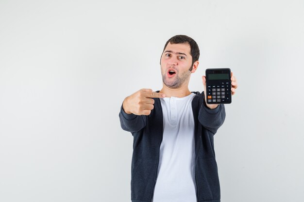 Jovem segurando calculadora e apontando para ela com o dedo indicador em uma camiseta branca e zíper frontal com capuz preto e olhando surpreso, vista frontal.