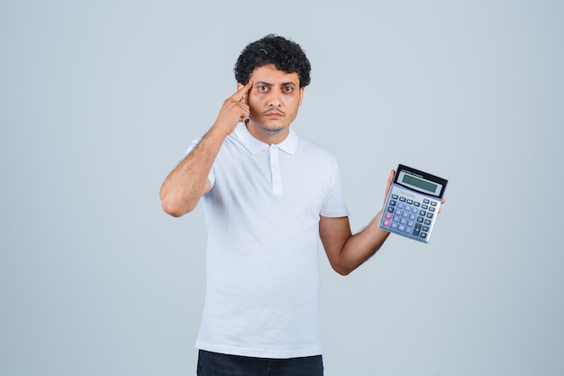 Jovem segurando calculadora, mantendo o dedo nas têmporas em t-shirt branca e parecendo inteligente. vista frontal.