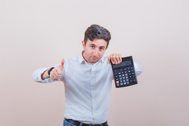 Jovem segurando calculadora, mostrando o polegar em uma camisa branca, jeans e parecendo satisfeito