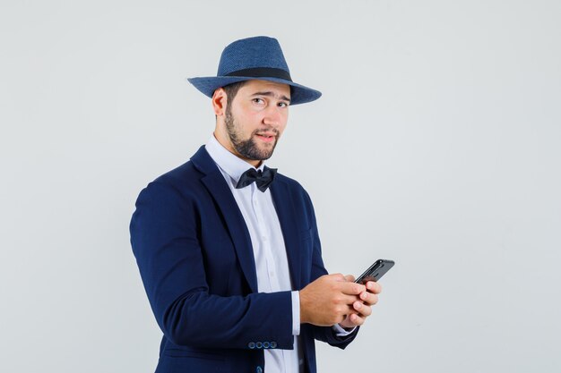 Foto grátis jovem, segurando o celular de terno, chapéu e olhando confiante, vista frontal.