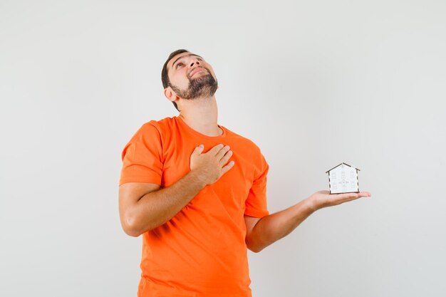 Jovem, segurando o modelo da casa com a mão no peito em t-shirt laranja e parecendo um sonho. vista frontal.