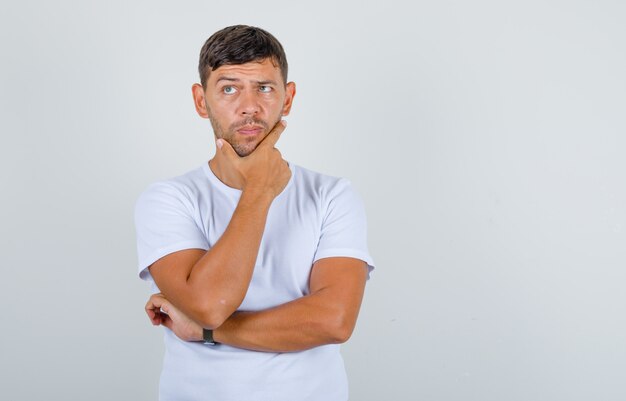 Jovem, segurando o queixo e levantando as sobrancelhas em t-shirt branca e olhando pensativo, vista frontal.