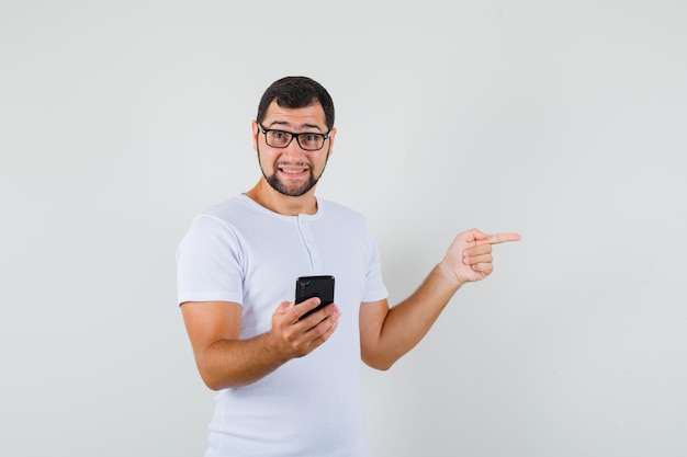 Jovem, segurando o telefone celular, apontando para o lado em uma camiseta branca, óculos e olhando animado, vista frontal.