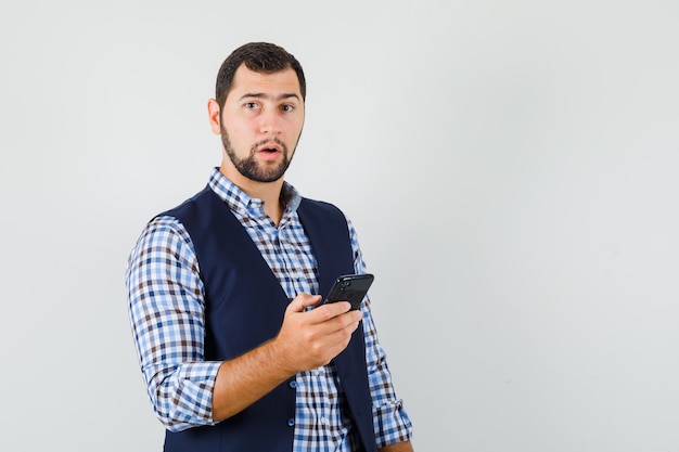 Jovem, segurando o telefone celular em uma camisa, colete e olhando pensativo.