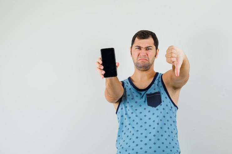 Foto grátis jovem, segurando o telefone celular, mostrando o polegar para baixo na camiseta azul e parecendo insatisfeito.