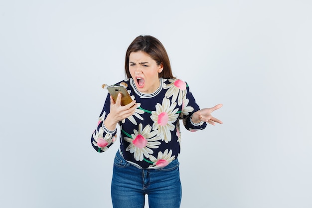 Foto grátis jovem, segurando o telefone, ficando descontente com a pergunta idiota numa blusa floral, jeans e parecendo ressentida, vista frontal.