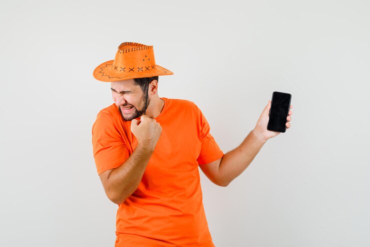 Foto grátis jovem, segurando o telefone móvel com o gesto de vencedor em t-shirt laranja, chapéu e olhando feliz, vista frontal.