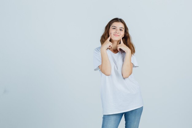 Jovem, segurando os dedos nas bochechas em t-shirt, jeans e linda. vista frontal.