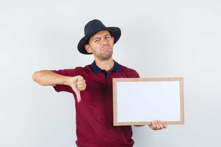 Foto grátis jovem segurando quadro branco com o polegar para baixo em t-shirt, chapéu e olhando descontente. vista frontal.
